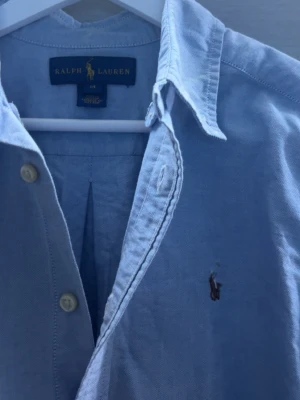 Ljusblå skjorta (Ralph Lauren) - Klassisk ljusblå skjorta från Ralph Lauren med broderad logga på bröstet. Skjortan har knappar framtill, krage och långa ärmar. Tillverkad i bomull för en bekväm känsla och snygg passform.