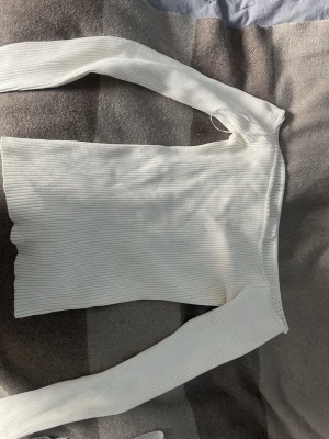Vit ribbad offshoulder topp - Trendig vit ribbad topp med offshoulder-design och långa ärmar. Sitter tight och har en snygg, figurnära passform som framhäver axlarna. Perfekt att styla med både jeans och kjol för en clean look.