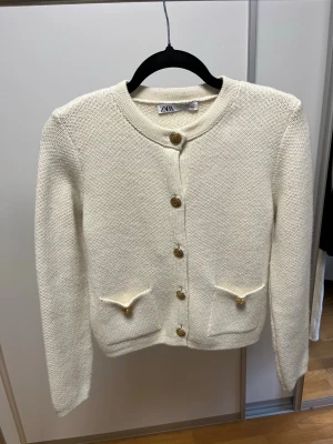 Krämvit kofta från Zara med guldknappar - Stilren krämvit kofta från Zara med rund halsringning och dekorativa guldknappar framtill. Koftan har två små fickor med lock och är stickad i ett mjukt material som känns lyxigt. Perfekt att slänga över en topp för en chic look.