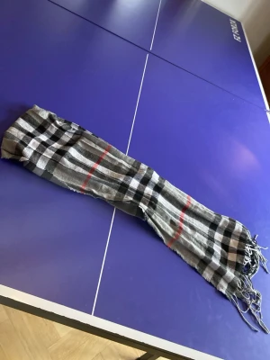 Rutig halsduk i grått och svart - Snygg halsduk med klassiskt rutmönster från Burberry i grått, svart, vitt och röda detaljer. Halsduken har fransar i ändarna och är tillverkad i ett mjukt tyg som känns skönt mot huden. Perfekt accessoar för att lyfta din outfit.