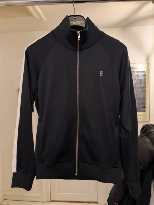 Svart träningsjacka från Björn Borg - Snygg svart träningsjacka från Björn Borg med vit rand längs ärmarna och dragkedja framtill. Jackan har hög krage, ribbade muddar och diskret B-logga på bröstet. 