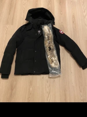 Canada Goose Wyndham. - Svart dunjacka från Canada Goose med huva och avtagbar pälskant. Klassisk patch på ärmen och flera praktiska fickor. Jackan är quiltad och har dragkedja framtill. Perfekt för kalla vinterdagar.