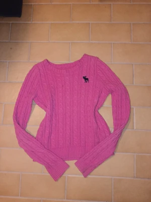 Rosa kabelstickad tröja från Abercrombie & Fitch - Rosa kabelstickad tröja från Abercrombie & Fitch med rund halsringning och svart älglogga broderad på bröstet. Tröjan har långa ärmar och ribbade muddar. Perfekt för lager-på-lager och en chill look.