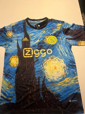 Ajax x Adidas Van Gogh tröja - Unik Ajax fotbollströja från Adidas med Van Gogh-inspirerat motiv i blått, gult och svart. Tröjan har Ajax-logga, Adidas-märke och Ziggo sponsortryck på bröstet. Tillverkad i lätt funktionsmaterial med Aeroready-teknologi.