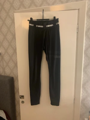 Svarta leggings från Stronger - Svarta leggings från Stronger med logotyp i vitt på midjeresåren. Hög midja och tight passform, tillverkade i stretchigt syntetmaterial. Diskret mönsterdetalj på ena benet. Perfekta för träning eller chill.