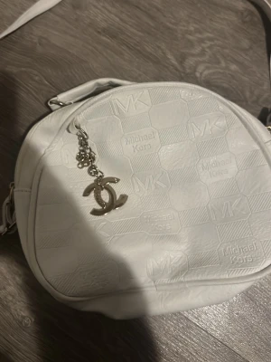 Vit axelväska Michael Kors - Snygg vit axelväska från Michael Kors med präglad logga över hela väskan. Väskan har en oval form, justerbar axelrem och ett stort fack med dragkedja. Utsidan är i konstläder och insidan har ett mönstrat tygfoder. Dekorerad med en silverfärgad nyckelring.