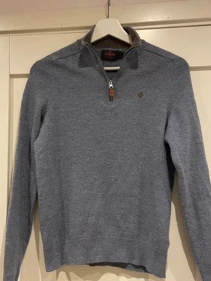 Ljusblå stickad tröja från Morris - Snygg ljusblå stickad tröja från Morris i 100% merinoull. Tröjan har halv dragkedja med läderdetalj, ståkrage och diskret broderad logga på bröstet. Perfekt för lager-på-lager och en stilren look.