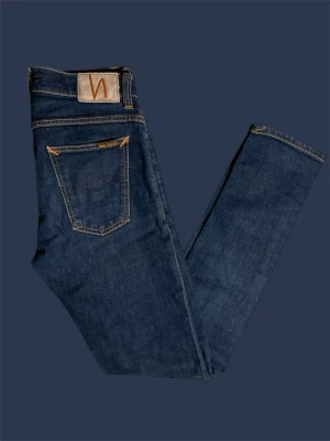 Mörkblå  jeans från Nudie - Säljer ett par mörkblå  jeans från Nudie Jeans med klassisk femficksdesign och orange kontrastsömmar. Jeansen har en smal passform och är tillverkade i mjuk denim med Nudie-logga på bakfickan. Perfekta för dig som gillar stilrena och tidlösa jeans.