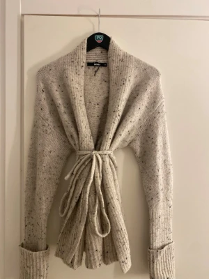 Ljusgrå stickad kofta med knytband - Så fin ljusgrå stickad kofta från Bik Bok med melerade detaljer och långa ärmar. Koftan har ribbstickade muddar och knytband i midjan för en snygg siluett. Nyskick💕