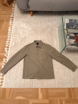 Olivgrön skjorta med bröstficka - Stilren olivgrön overshirt med klassisk krage och knappstängning framtill. Skjortan har en bröstficka och långa ärmar. Dessvärre ett litet hål längst ner på höger ärm. Därav det låga men bra priset!
