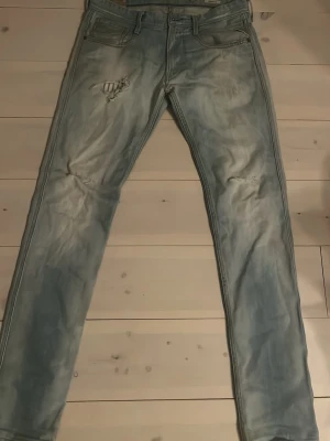 Ljusblå Replay jeans med slitningar - Säljer ett par ljusblå jeans från Replay med snygga slitningar och tvättad look. Jeansen har klassisk femficksmodell och raka ben. Perfekta för dig som gillar en avslappnad och trendig stil.
