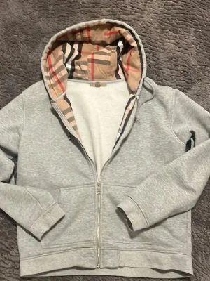 Grå hoodie med Burberry-mönstrad huva - Snygg grå zip-hoodie från Burberry med den klassiska rutiga insidan av huvan. Mjuk bomullskvalitet, dragkedja framtill och ribbade muddar. Perfekt för dig som vill ha en stilren hoodie med lyxig touch. Storlek xl men ganska liten i storlek, passar L