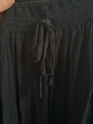 Svart långkjol med snörning - Svart långkjol med resår och snörning i midjan. Kjolen är tillverkad i ett lätt, skrynkligt tyg som ger en avslappnad känsla. Perfekt att styla med en enkel topp eller hoodie.