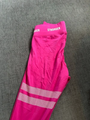 Rosa träningstights från Stronger - Rosa träningstights från Stronger med hög midja och vita logotyper i midjan. Tightsen har två vita randiga detaljer med prickmönster på ena benet och är tillverkade i stretchigt syntetmaterial. Inga tecken på användning💕