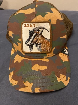 Camo truckerkeps med get-brodyr - Säljer en camouflagemönstrad truckerkeps från Goorin Bros med mesh baktill och justerbar snapback. Framtill finns en stor patch med en broderad get och texten 'GOAT'. Perfekt för dig som gillar streetwear och djurmotiv. Trasigt spänne funkar på några.