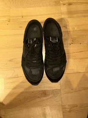 Svarta Valentino sneakers med nitar - Snygga svarta sneakers från Valentino med mattsvarta detaljer och coola nitar på hälen. Skorna har en låg profil, svarta skosnören och en blandning av läder och textilmaterial. Perfekta för dig som gillar en stilren och edgy look. Skolåda dustbag och annat finns har små skador som inte syns så bra där av har dom ett billigare pris 
