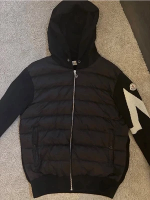 Svart dunjacka med huva från Moncler - Snygg svart dunjacka från Moncler med huva och dragkedja. Ena ärmen har en vit zigzag-detalj och Moncler-logga. Jackan har två fickor framtill och quiltad front. Perfekt för kalla vinterdagar.