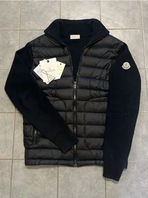 Svart pufferjacka från Moncler - Snygg svart pufferjacka från Moncler med quiltad front och stickade ärmar. Jackan har dragkedja framtill, ståkrage och två sidofickor med dragkedja. Moncler-logga på vänster ärm. Perfekt för kalla dagar.
