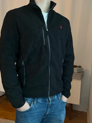 Svart Ralph Lauren fleece, storlek M - Svart Ralph Lauren fleece med röd logga och grå dragkedjor.   100% polyester  Mycket bra skick, inga defekter, jättefräsch   Storlek M Axelbredd: 48 cm Längd: 69 cm