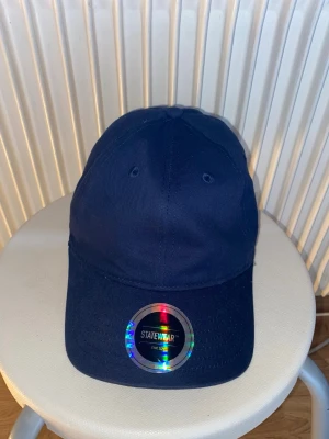 Mörkblå keps från Statewear - Snygg mörkblå keps från Statewear med klassisk böjd skärm och justerbart spänne bak. Tillverkad i slitstark bomull och har ventilationshål för extra komfort. Perfekt accessoar för en avslappnad streetwear-look.