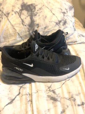 Nike Air Max 270 svarta sneakers - Säljer ett par svarta Nike Air Max 270 sneakers med vit sula och synlig Air-enhet i hälen. Skorna har mesh-ovandel för bra ventilation, klassisk Nike Swoosh på sidan och AIR270-tryck. Perfekta för dig som gillar sportig och modern stil.