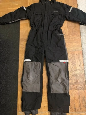 Svart täckoverall Didriksons 140 - Täckoverall från Didriksons i svart med grå förstärkningar på knäna och reflexdetaljer. Overallen har dragkedja, fickor och elastisk midja. Insidan är fodrad med gul textil. Perfekt för kalla vinterdagar och utomhusaktiviteter.