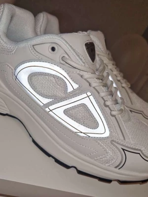 Vita Dior B30 sneakers med reflexdetaljer - Snygga vita Dior B30 sneakers med reflekterande detaljer på sidorna och svart sula. Ovandelen är i mesh och syntetmaterial för en modern look. Klassisk snörning och tydlig Dior-logga på plösen. Perfekta för dig som vill sticka ut med stil.