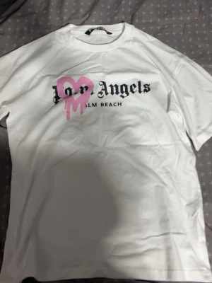 Vit Palm Angels t-shirt med tryck - Vit t-shirt från Palm Angels med svart text och ett stort rosa hjärta med drip-effekt på bröstet. Klassisk passform och rund halsringning. Perfekt för dig som gillar streetwear och vill sticka ut med en unik design.