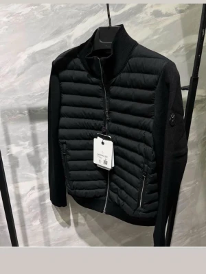Svart cardiganjacka från Moncler - Snygg svart cardiganjacka från Moncler med quiltad front och stickade ärmar. Jackan har hög krage, två dragkedjefickor och Moncler-logga på ärmen. Perfekt för lager på lager och ger en clean look. Materialmix av dun och ull för både värme och stil.