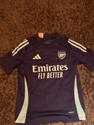 Arsenal träningströja från Adidas - Mörklila Arsenal träningströja från Adidas med vita detaljer och logga på bröstet. Tröjan har trycket 'Emirates Fly Better' på framsidan och klassiska Adidas-ränder på axlarna. Tillverkad i funktionsmaterial för sport.