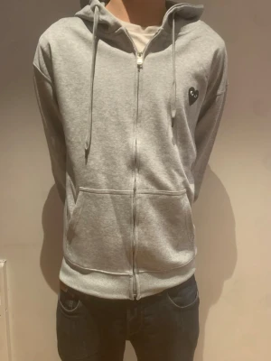 Grå Comme des Garçons Play hoodie - Pris går att diskuteras!!! Grå zip-hoodie från Comme des Garçons Play med svart hjärta och ögon-tryck på ryggen samt litet hjärta på bröstet. Mjuk bomull, dragsko i huvan och två fickor framtill. Perfekt för en chill och stilren look.