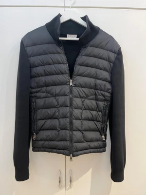 Svart pufferjacka från Moncler - Svart jacka från Moncler. Vadderad framtill och stickade ärmar och rygg. Dragkedja fram och två fickor med dragkedja. Moncler-märke på ärmen.