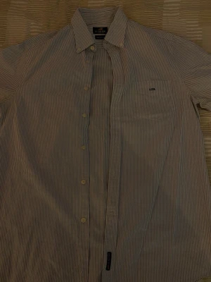 Randig skjorta från U.S. Polo Assn. - Klassisk skjorta med smala blå och vita ränder från U.S. Polo Assn. Skjortan har knappar framtill, bröstficka med broderad logga och krage. Tillverkad i bomull för en skön känsla.