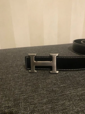 Svart skärp med H-spänne - Stilrent svart skärp med ett ikoniskt H-format spänne i silverfärgad metall. Skärpet har vita kontrastsömmar längs kanterna. Perfekt accessoar för att lyfta din outfit.