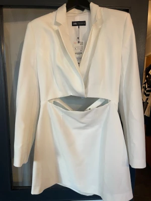 Vit cut out klänning från Zara - Stilren vit klänning från Zara med cut out-detalj i midjan och bälte med spänne. Klänningen har lång ärm, kavajkrage och kort längd. Perfekt för dig som vill sticka ut med en modern look.