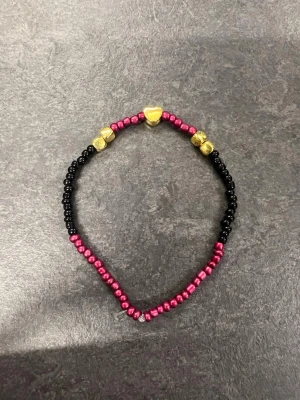 Armband i svart, röd och guld storlek L  - Säljer ett handgjort armband med svarta och röda pärlor samt detaljer i guldfärg, inklusive en hjärtformad pärla. 