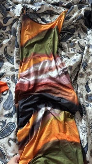 Färgglad långklänning med smala axelband - Säljer en långklänning med smala axelband och färgglada horisontella ränder i orange, grön, svart, vit och vinröd. Klänningen har en figurnära passform och är tillverkad i ett mjukt, stretchigt material som känns bekvämt mot huden.