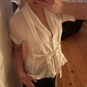 Vit blus - Vit blus med knappar framtill och knyte i midjan. Knytet går att välja vart man vill knyta, som man kan se på bilderna. Har rosettdetaljer på sidan av ärmarna! 🌷