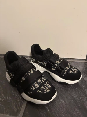 Svarta Dior sneakers med monogram - Snygga svarta sneakers från Dior med tydligt monogrammönster i vitt och svart. Skorna har breda kardborreband, meshdetaljer och en chunky vit sula med svart stjärndetalj undertill. Perfekta för dig som gillar statement-skor och exklusiv streetstyle.