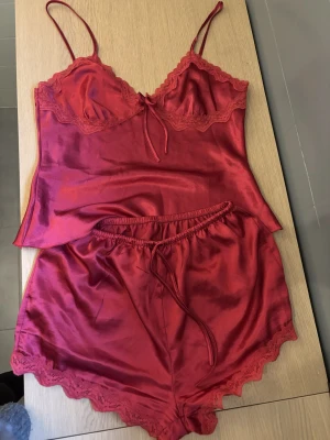 Rött satinsovset med spetsdetaljer - Snyggt rött sovset i satin med linne och shorts. Linnet har tunna axelband, spetskant och liten rosett framtill. Shortsen har elastisk midja och spetskant nedtill. Perfekt för dig som gillar mjuka material och en lyxig känsla.