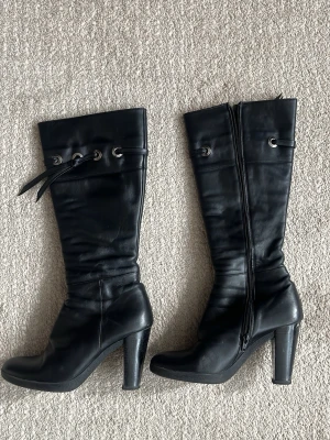 Svarta höga stövletter med snörning - Snygga svarta stövletter i skinnimitation med högt skaft och grov klack. Dekorativ snörning med metallöljetter upptill och dragkedja på insidan. Perfekta för att lyfta din outfit med en edgy touch.