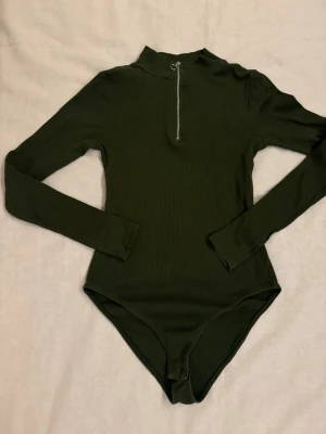 Mörkgrön ribbad body med dragkedja - Snygg mörkgrön body med lång ärm och ribbad struktur. Hög krage och silverfärgad dragkedja framtill som detalj. Perfekt att styla med jeans eller kjol. Materialet känns mjukt och stretchigt.