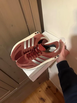 Adidas L.A Trainer - sneakers i vinröd mocka - Säljer ett par snygga Adidas sneakers i vinröd mocka och mesh med klassiska vita ränder på sidorna. Modell: la trainer. Skorna har en retroinspirerad design, vita sulor och röda skosnören.