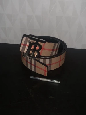 Beige rutigt bälte från Burberry - Snyggt bälte från Burberry i beige med klassiskt rutig design i rött, svart och grått. Bältet har ett stort metallspänne med ett B i silverfärg. Perfekt för att lyfta din outfit med en ikonisk accessoar.                           110cm