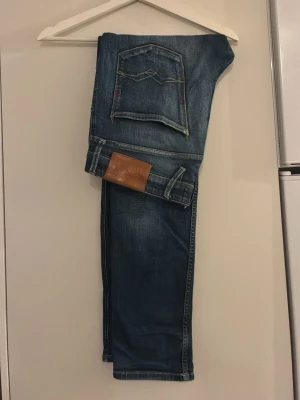 Blå replay jeans med raka ben - Säljer ett par klassiska blå replay jeans med raka ben och femficksmodell. Jeansen har en snygg tvättad look, kontrastsömmar och ett brunt läderpatch bak i midjan. Perfekta för en avslappnad och stilren outfit.