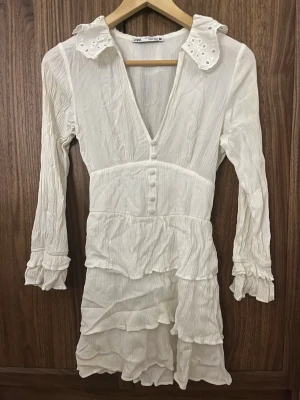Vit volangklänning från Zara - Supersöt vit kortklänning från Zara med volanger, v-ringning och dekorativa knappar framtill. Klänningen har långa ärmar med volangdetaljer och en krage med broderade hål. Perfekt för dig som gillar romantisk och trendig stil.