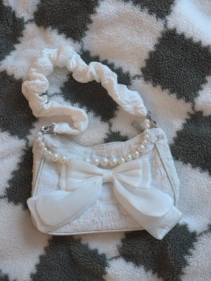 Vit handväska med rosett och pärlor - Supersöt vit handväska med stor rosett framtill och dekorativa pärlor längs ovankanten. Väskan har ett scrunchie-inspirerat axelband och är gjord i ett tyg med struktur. Dragkedja upptill och ljus insida. Perfekt för dig som gillar gulliga detaljer och unik design.