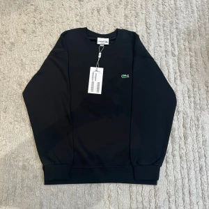 Svart sweatshirt från Lacoste - Svart sweatshirt från Lacoste med klassisk rund halsringning och det ikoniska gröna krokodilmärket broderat på bröstet. Tröjan har ribbade muddar vid ärmslut och nederkant. Perfekt för en stilren och avslappnad look.