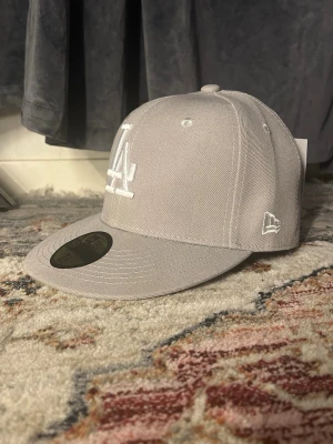 Grå LA-keps från New Era - Snygg grå keps från New Era med LA-logga broderad framtill i vitt och World Series-märke på sidan. Klassisk flat peak och strukturerad passform. Perfekt accessoar för en sportig och trendig look.