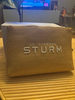 Beige necessär Dr. Barbara Sturm - Snygg beige necessär från Dr. Barbara Sturm i grovt tyg med vit broderad logga på framsidan. Väskan har dragkedja och insidan är fodrad i gult tyg med tryckt logga. Perfekt för småsaker eller smink.